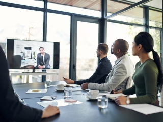 Four people sitting around an meeting table, looking at one person on a screen.